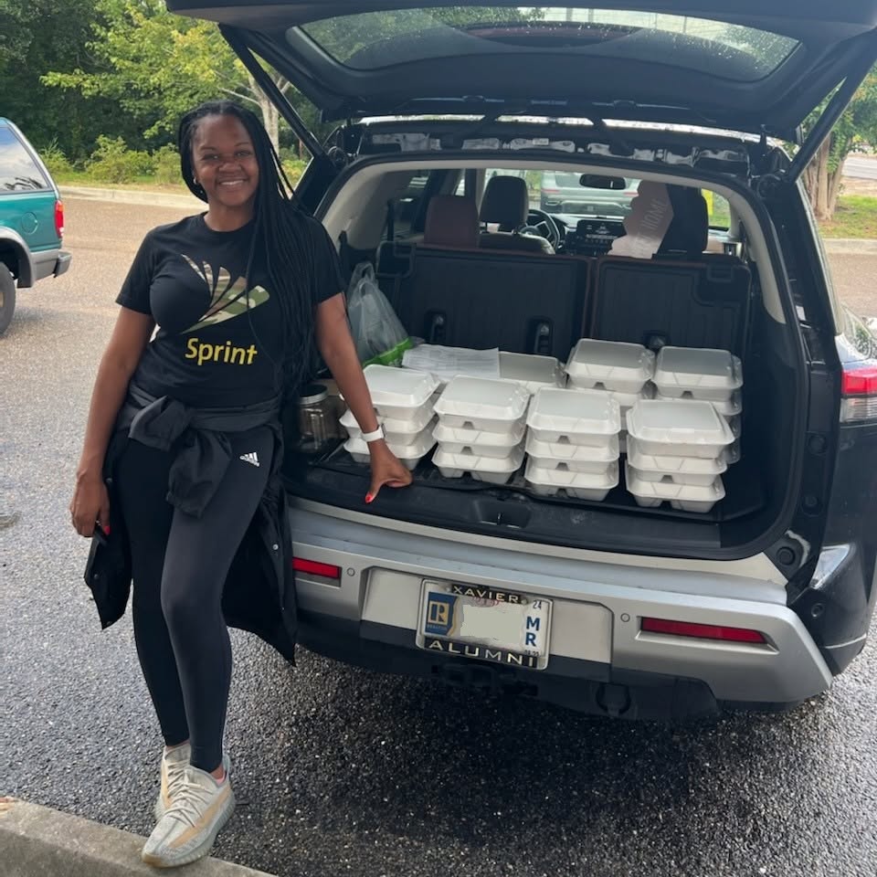 A smiling volunteer beside an SUV loaded with boxed hot meals ready for a Meals on Wheels route